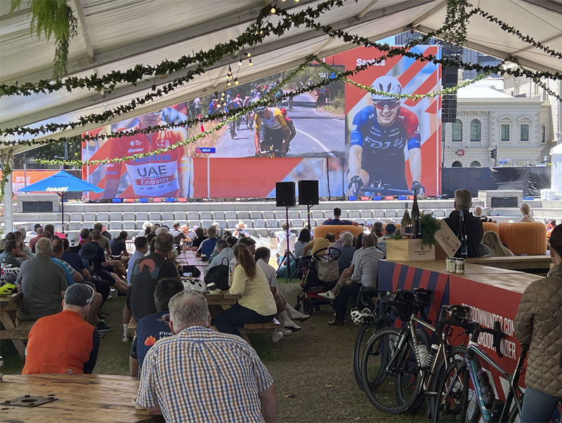 Spectators watching bicycle race on large screen under pavilion