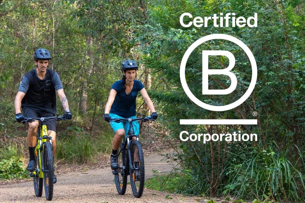 Two people riding bikes on bush track. B Corporation Certification logo appears to the right.