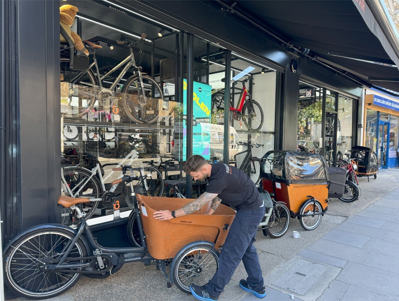 Man positioning cargo bike onto street in front of shop for display.