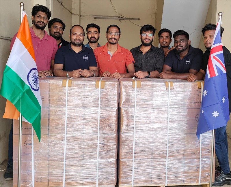 Group of men standing with large boxes. Indian and Australian flags stand to either side.